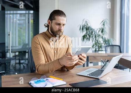Ein professioneller Mann sitzt an einem Schreibtisch in einem modernen Büro, hält ein Tablet und konzentriert sich auf die Arbeit. Er verfügt über ein Notebook, ein Notebook und Zubehör, das auf die Produktivität hinweist. Stockfoto