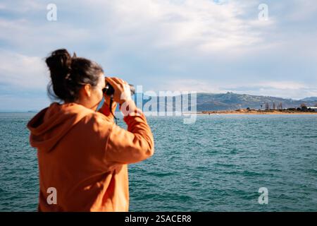 Eine junge Frau mit einem orangefarbenen Hoodie wird von hinten gesehen und mit einem Fernglas die malerische Küste von Santoña in Kantabrien, Spanien, bewundert. Das tiefe blu Stockfoto