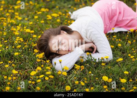 Junge Frau, die auf einem grasbewachsenen Feld liegt, bedeckt mit gelben Löwenzahn und genießt den Frühling. Stockfoto