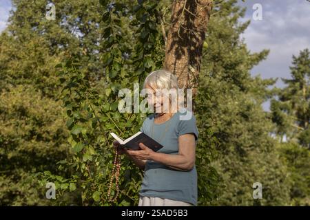 Ältere Frau, die in einem Garten steht und die Bibel liest und Gebetskegeln hält Stockfoto