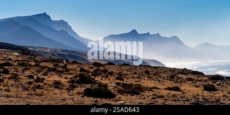 Ein Panoramablick auf zerklüftete Berge und eine Küste unter einem klaren blauen Himmel. Der Vorderboden weist trockenes, felsiges Gelände mit karger Vegetation auf Stockfoto