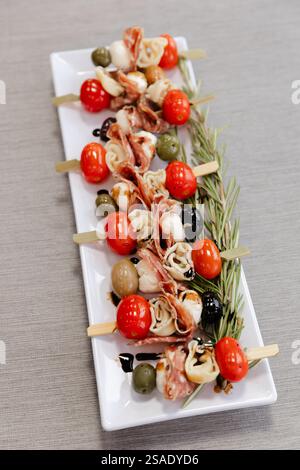 Farbenfrohe Spieße zeigen eine Mischung aus gegrillten Kirschtomaten, Oliven und in Fleisch gewickeltem Käse, die ordentlich auf einer weißen Platte in einem modernen Speisesaal angeordnet sind Stockfoto
