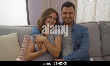 Mann und Frau sitzen zusammen auf einer Couch in einem gemütlichen Wohnzimmer drinnen, lächeln und umarmen einander warm. Stockfoto