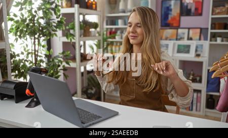 Frau, die sich im Innenraum des Wohndekors mit Laptop mit Schürze und Lächeln entspannt, während sie Brille mit stilvollem Hintergrund von dekorativen Gegenständen hält und lächelt Stockfoto