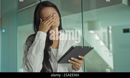 Frau, die sich gestresst fühlt und im Büro eine Tablette hält, die junge chinesische Frau mit Hand auf Gesicht zeigt Stockfoto
