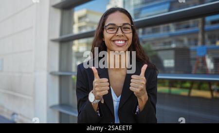 Eine junge Frau mit einer Brille, die lächelt und die Daumen nach oben zeigt, in einer urbanen Umgebung, in einem schwarzen Blazer vor einer Straßenkulisse mit Gebäuden Stockfoto
