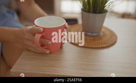 Frau hält Kaffeetasse in gemütlichem Café-Setting mit Pflanzen- und Korbuntersetzer auf Holztisch Stockfoto