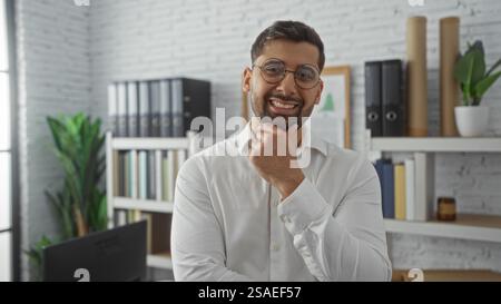 Junger Mann, der selbstbewusst lächelt in moderner Büroumgebung mit Bücherregalen und natürlichem Licht, das ein professionelles und dennoch ansprechendes Ambiente widerspiegelt Stockfoto