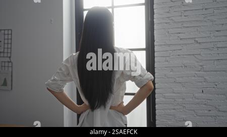 Attraktive junge Frau mit langen Haaren steht drinnen in einem Bürozimmer und blickt nachdenklich aus dem Fenster mit Händen auf den Hüften gegen einen weißen Ziegelstein Stockfoto