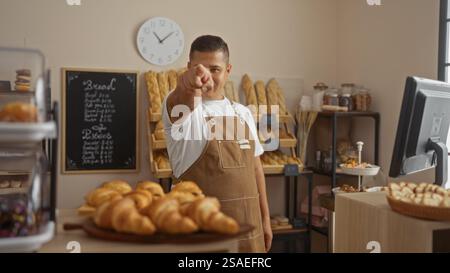 Junger hispanischer Mann, der in einer Bäckerei in Richtung der Kamera zeigt, in einer Innenumgebung, gefüllt mit verschiedenen Brotsorten und Gebäck Stockfoto