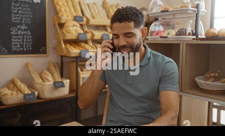 Junger Mann, der telefoniert, während er in einer gemütlichen Bäckerei sitzt, umgeben von Brot und Gebäck Stockfoto