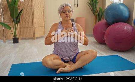 Reife Frau, die Yoga in einem Fitnessstudio praktiziert, mit geschlossenen Augen und Händen auf der Brust, umgeben von Übungsbällen und Pflanzen. Stockfoto