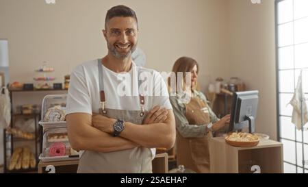 Mann lächelt mit überkreuzten Armen und Frau arbeitet an einem Computer im Hintergrund in einer Bäckerei, die Teamarbeit und eine freundliche Atmosphäre zeigt. Stockfoto