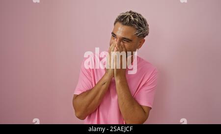 Junger arabischer Mann mit Bart und Tätowierung lacht, während er sein Gesicht bedeckt, mit rosa Hemd isoliert über rosa Hintergrund Stockfoto