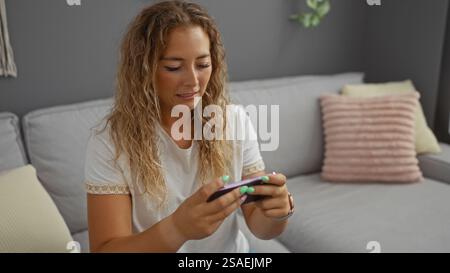 Schöne blonde Frau, die auf einem bequemen Sofa in einem gemütlichen Wohnzimmerapartment ein Smartphone spielt Stockfoto
