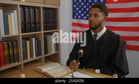 Junger afroamerikanischer Mann, der Richterroben trägt und im Gerichtssaal einen Hammer hält, mit amerikanischer Flagge im Hintergrund und Bücherregalen voller Rechtstexte. Stockfoto