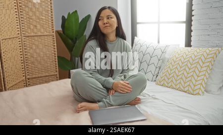 Junge Frau, die auf dem Bett in einem gemütlichen Schlafzimmer sitzt, bequeme Loungewear trägt, mit Laptop, genießt einen friedlichen Morgen drinnen und entspannt sich zu Hause in einem Ser Stockfoto