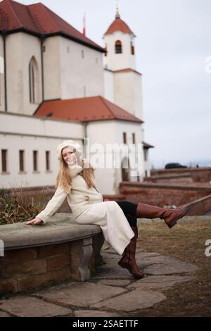 Eine modische Frau in beigefarbenem Winteroutfit steht an einer historischen Backsteinmauer in der Nähe eines Schlosses Spilberk in Tschechien. Gemütliche und elegante Atmosphäre. Stockfoto