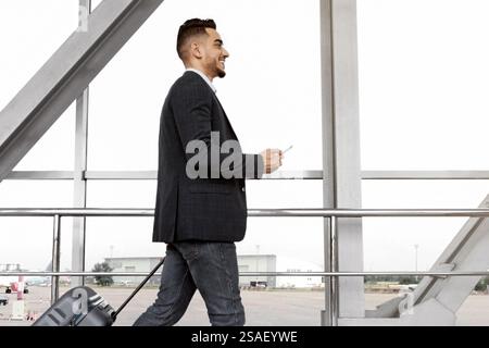 Junger Geschäftsmann aus dem Nahen Osten mit Koffer und Smartphone in den Händen, der auf dem Flughafen unterwegs ist Stockfoto