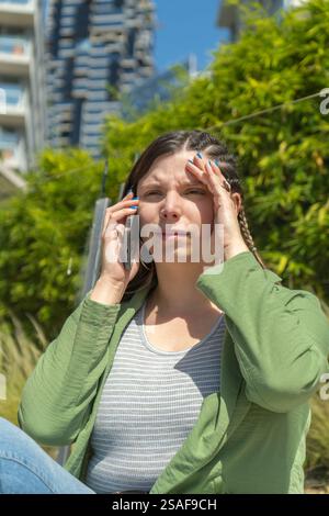 Gestresste junge Frau, die Kopfschmerzen beim Telefonieren in einem Stadtpark hat Stockfoto