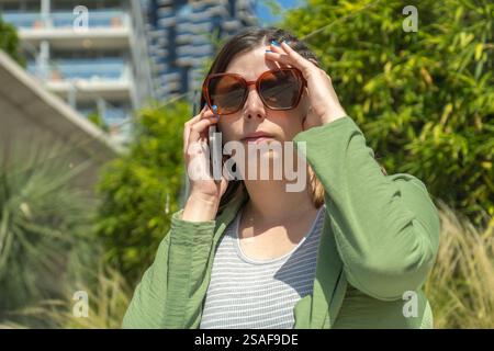 Eine junge Frau, die auf einem Smartphone spricht und ihre Augen vor der Sonne schützt Stockfoto