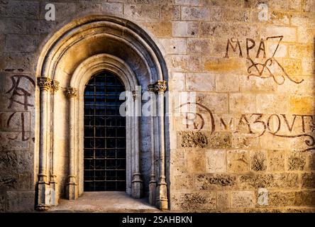 Eine historische Steinmauer mit einem bogenförmigen Fenster und einem schmiedeeisernen Grill. Die Wand ist mit antiken Inschriften und Graffiti geschmückt, die einen zeigen Stockfoto