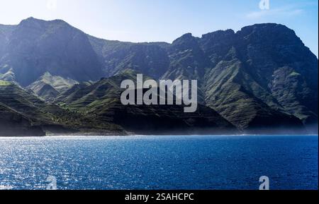 Ein malerischer Blick auf zerklüftete Berge, die steil aus dem Meer ragen, mit üppiger grüner Vegetation an den Hängen und einem klaren blauen Himmel darüber. Das Sonnenlicht cas Stockfoto
