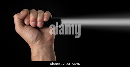 Schwarze Taschenlampe in menschlichen Händen auf schwarzem Hintergrund, einschließlich eines weißen Strahls. Stockfoto