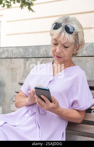 Eine ältere Frau im Lavendelkleid sitzt auf einer Bank und benutzt ein Smartphone Stockfoto
