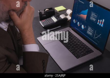 Ein Geschäftsmann in einem Anzug analysiert Finanzdaten auf einem Laptop und überlegt Anlagestrategien. Ein Taschenrechner, Notizen und Büromaterial erstellen einen Stockfoto