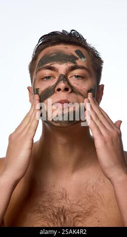 Junger Mann mit nassen, brünetten Haaren, der Hautpflegeverfahren durchführt, Nährtonmaske auf der Haut vor weißem Hintergrund aufträgt. Stockfoto