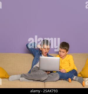 Schockierter Junge, der mit Bruder über Laptop Film sieht, während er auf dem Sofa vor violettem Hintergrund sitzt Stockfoto