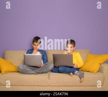 Lächelnde Brüder, die sich Filme ansehen und Videospiele über Laptops auf dem Sofa vor violettem Hintergrund spielen Stockfoto