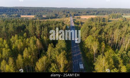Aus der Vogelperspektive auf einer geraden Straße, die durch einen dichten Wald führt, auf dem Militärkonvoi unterwegs sind, umgeben von üppigen grünen Bäumen und Feldern unter einem Stockfoto