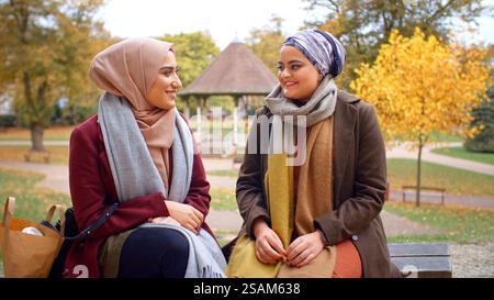 Zwei Junge Britische Muslimische Freundinnen Sitzen Auf Der Bank Im Autumn Park Stockfoto