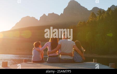 Rückansicht Der Liebevollen Familie, Die Auf Einem Holzsteg Am See Bei Sonnenuntergang Mit Lens Flare Sitzt Stockfoto