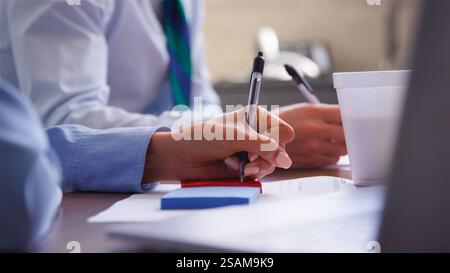 Nahaufnahme Der Besprechung Des Business-Teams, Die Sich Um Den Tisch Im Büro Herum Mit Getränketassen Zusammensetzt, Um Notizen Zu Machen Stockfoto