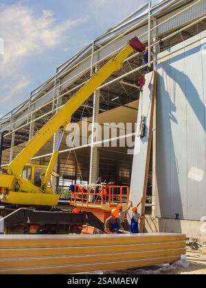 Installation von Sandwichpaneelen auf einem Metallgerüst eines Kühllagers unter Verwendung von Kränen und Liften. Stockfoto