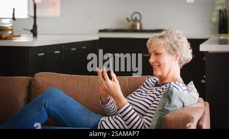 Ältere Frau Auf Sofa Zu Hause, Die Ihr Mobiltelefon Benutzt, Um Online-Streaming-Sendungen Zu Durchsuchen Oder Nachrichten Zu Lesen Stockfoto