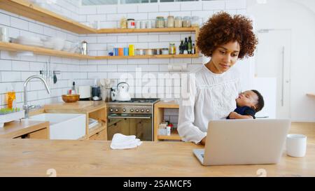 Multitasking-Mutter Hält Den Schlafenden Sohn, Während Sie In Der Küche An Einem Laptop Arbeitet Stockfoto