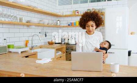 Multitasking-Mutter Hält Den Schlafenden Sohn, Während Sie In Der Küche An Einem Laptop Arbeitet Stockfoto
