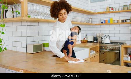 Multi-Tasking Mutter Hält Schlafenden Sohn Während Der Reinigung In Der Küche Zu Hause Stockfoto