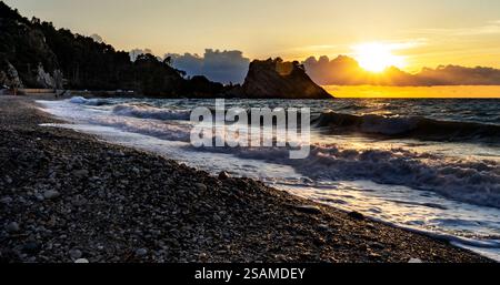 Eine ruhige Strandszene bei Sonnenuntergang mit sanften Wellen, die gegen ein Kieselstrand plätschern. Die Sonne untergeht hinter einem Felsvorsprung und erzeugt warme Farbtöne Stockfoto