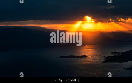 Ein dramatischer Sonnenuntergang über einem ruhigen Meer mit Sonnenstrahlen, die durch dunkle Wolken brechen und das Wasser und die fernen Berge beleuchten. Stockfoto