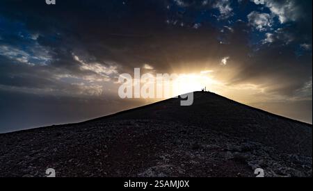 Ein dramatischer Sonnenuntergang über einem Berggipfel, mit Sonnenstrahlen, die durch dunkle Wolken brechen. Silhouettenfiguren stehen auf dem Gipfel und erzeugen einen Striki Stockfoto