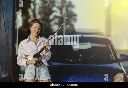 Eine Frau, die einen Hochdruckreiniger hält, steht in der Nähe eines blauen Autos und lächelt in der Sonne Stockfoto