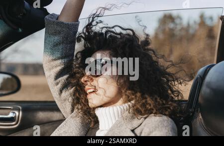 Fröhliche Frau mit Sonnenbrille in einem Cabrio, vom Wind geweht Haare, umgeben von Land Stockfoto