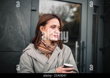 Frau in hellem Mantel und gestreiftem Schal, die eine Tasse hält, nachdenklich wirkt Stockfoto