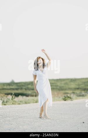 Lächelnde Frau in weißem Kleid, die fröhlich den Arm auf einem Weg erhebt, umgeben von einem Feld Stockfoto