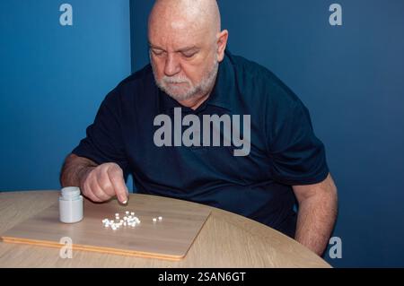 Ein kahlköpfiger Mann mittleren Alters, wahrscheinlich 50 oder 60, sitzt an einem Tisch und gießt Pillen aus einer weißen Flasche auf den Tisch. Das Bild schlägt eine bestellung vor Stockfoto